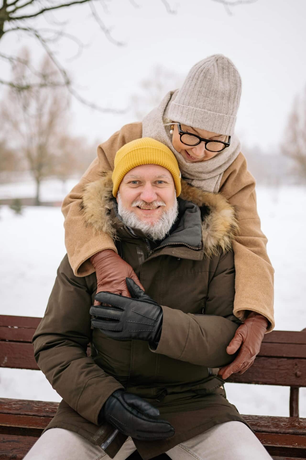 A man and woman in the snow sitting on a bench posing for the camera.